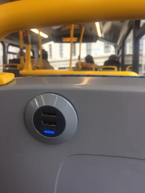 A close-up shot of the charging port of an electric school bus being plugged in. Focus on the charging cable and the modern design of the charging station, with a school building visible in the background, showing the scale of the vehicles and their proximity to a school.